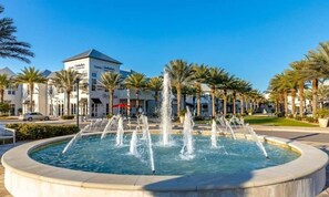 A circular fountain with multiple water jets in the center, surrounded by palm trees and a building with signage.