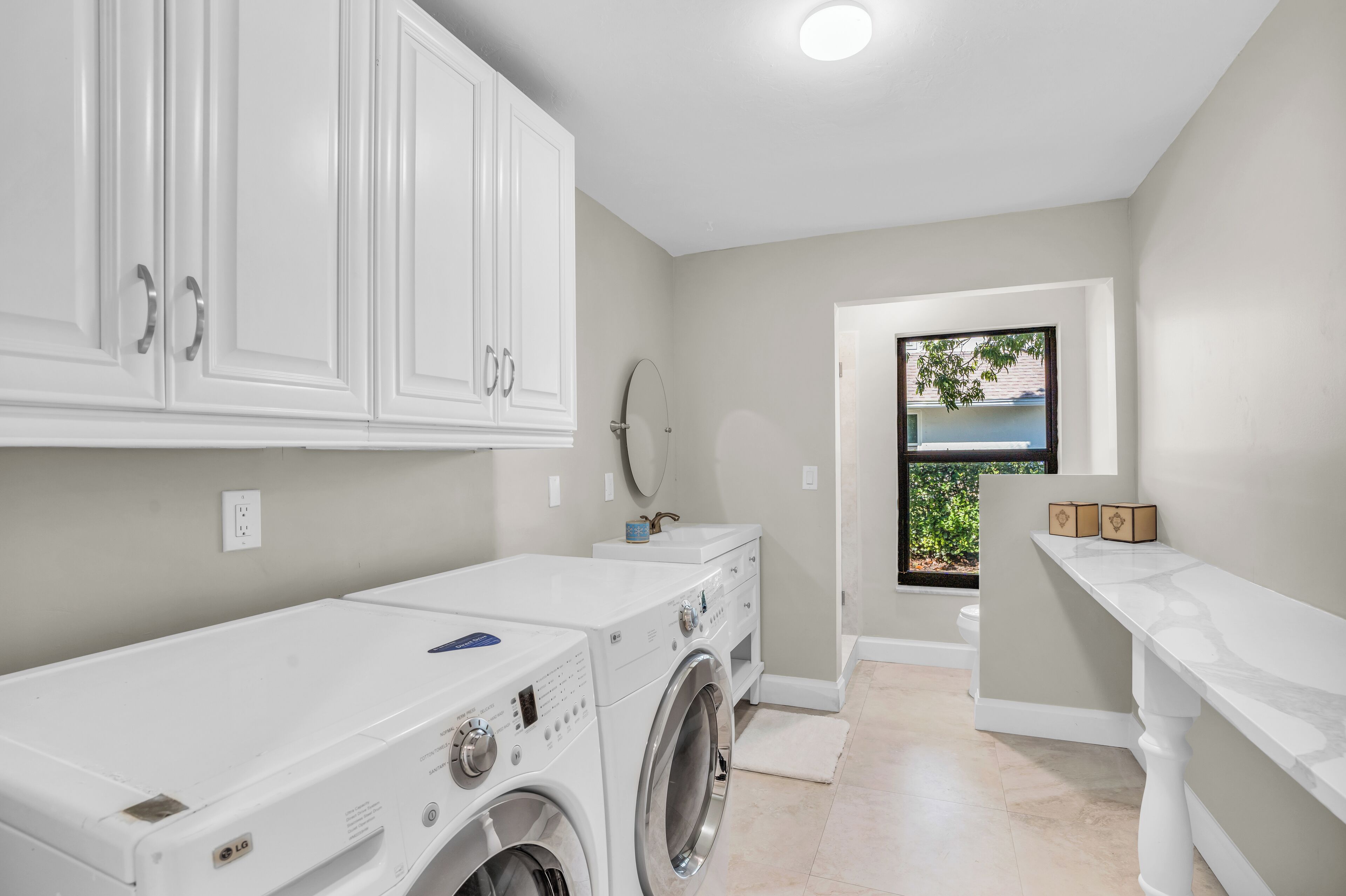 A laundry room with a front-loading washer and dryer, white cabinetry, and a window with a view of greenery.