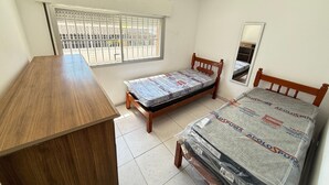 Een kleine slaapkamer met een houten bureau, een enkel bed met matras, een raam met metalen tralies en een spiegel aan de muur.