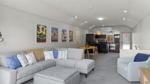 A modern living room with a grey sectional sofa, a white armchair, and a dining area with a wooden table and chairs. - Alpine Village 23 - 2 bed with lake views (Queenstown)