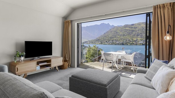 A modern living room with a flat-screen TV, a sofa, and a balcony with a view of a lake and mountains. - Alpine Village 23 - 2 bed with lake views (Queenstown)