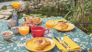 A table set with breakfast items including croissants, fruit, and orange juice, with a vase of flowers and a decorative bowl in the background. - ÔVILLAGE  (Ouveillan)