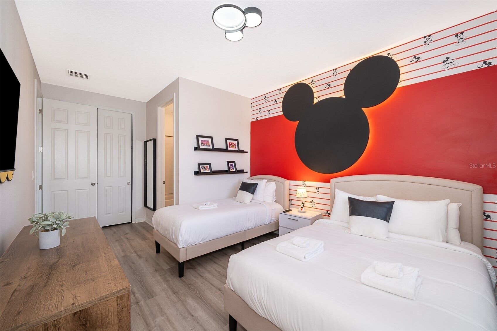 A hotel room with two beds, a wooden table with a plant, a Mickey Mouse mural, and framed pictures on the wall.