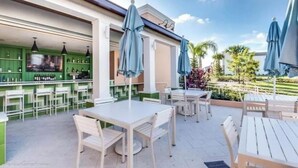Un patio al aire libre con mesas y sillas blancas, zona de bar verde y sombrillas.