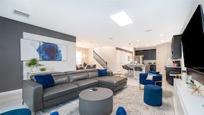 Una sala moderna con un sofá seccional gris, una mesa redonda de centro y sillones de acento azules. Una escalera conduce a una zona de cocina con un comedor.