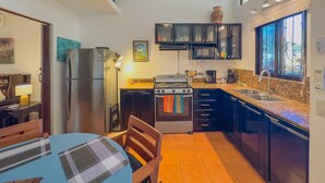 Een kleine keuken met een koelkast, kookplaat, magnetron en spoelbak. Er staat een eettafel met stoelen en een raam zorgt voor natuurlijk licht.