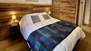 A bedroom with a wooden headboard, a bed with white bedding and patterned pillows, a blue and black checkered blanket, a nightstand with a lamp, and a framed picture on the wall. - Large, bright T4 in the heart of Châtel (CHATEL)