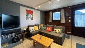 A cozy living room with a flat-screen TV, a sofa set, a coffee table, and a bookshelf with DVDs.