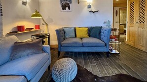 Ein gemütliches Wohnzimmer mit grauem Sofa, einem kleinen Tisch und einer Lampe. Es gibt Wandleuchten und ein Bärgemälde.