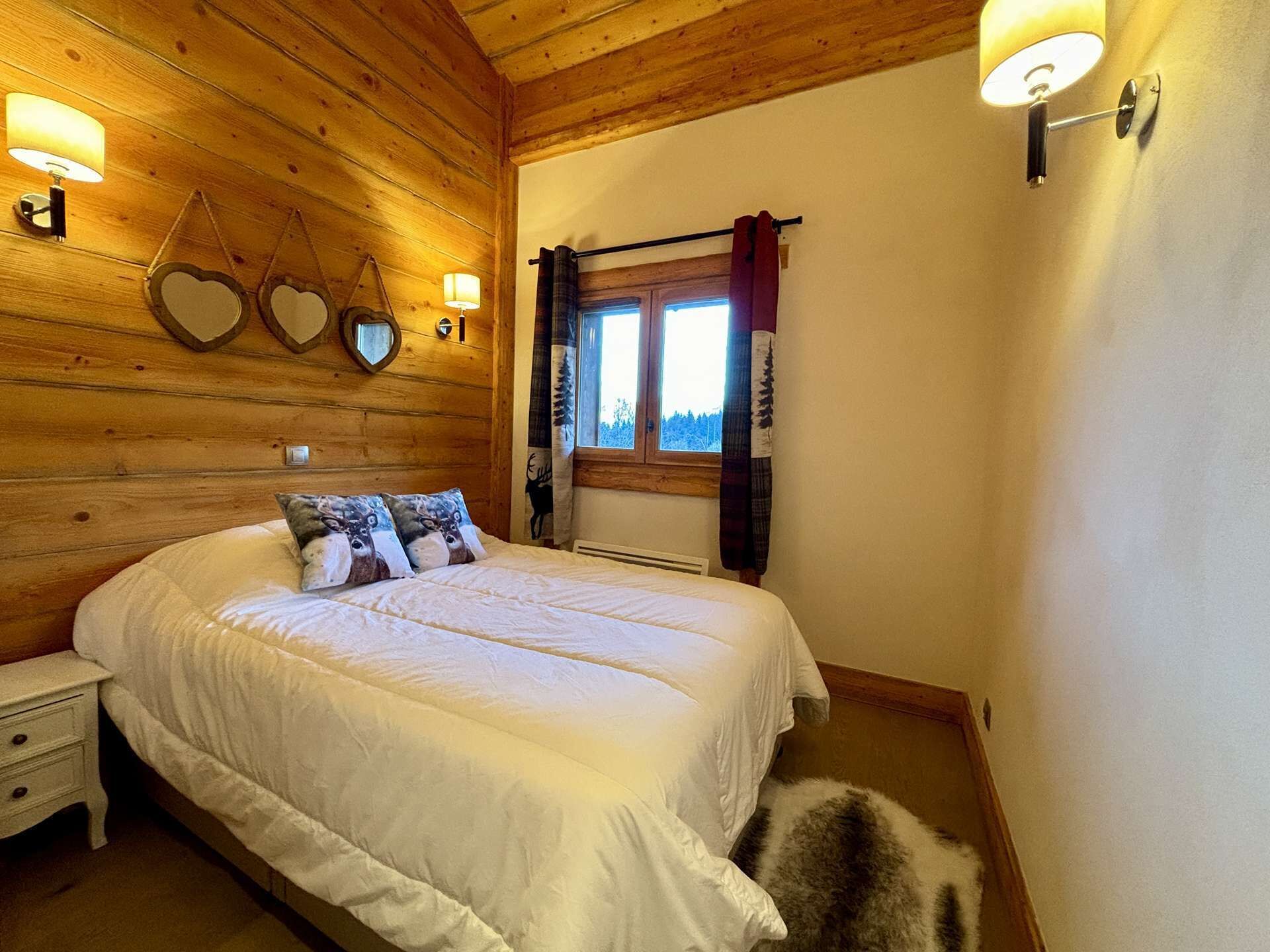 A cozy bedroom with a wooden ceiling, a bed with white bedding, heart-shaped wall decorations, and a window with curtains.