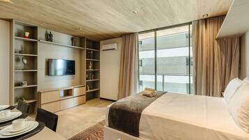 Habitación de hotel moderna con cama, televisor, zona de comedor y un gran ventanal con cortinas.