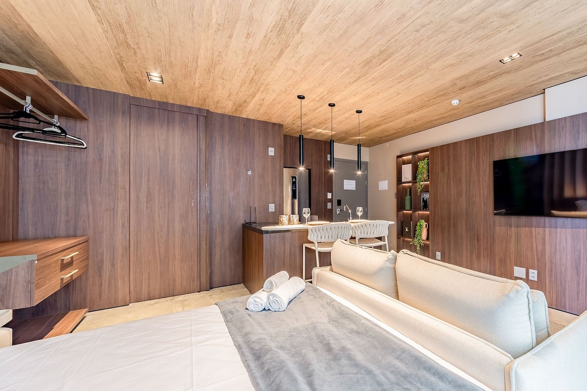 Una habitación de hotel moderna con techo de madera, un televisor de pantalla plana montado en la pared, una cama con cabecera gris y una zona de estar con sofá y una mesita.