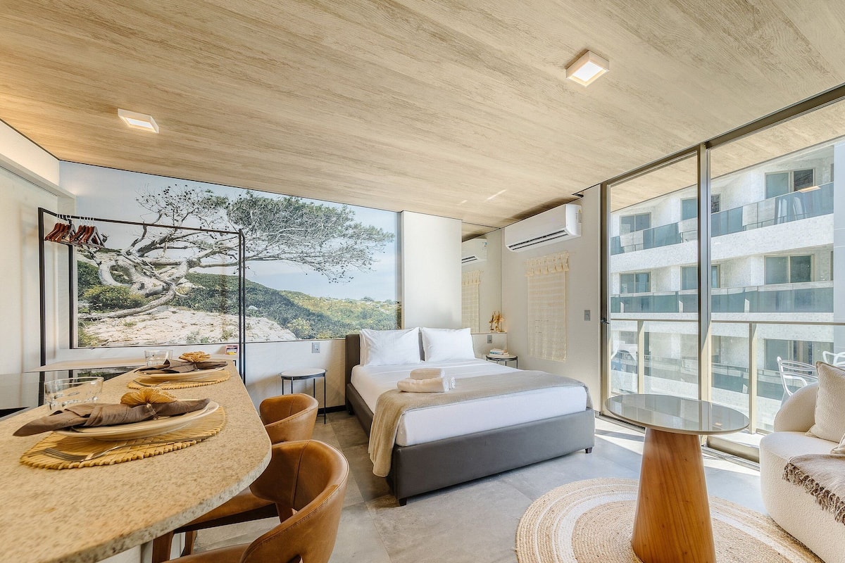Une chambre moderne avec un grand lit, un coin repas comprenant une table et des chaises, et une vue sur un immeuble à travers de grandes fenêtres.