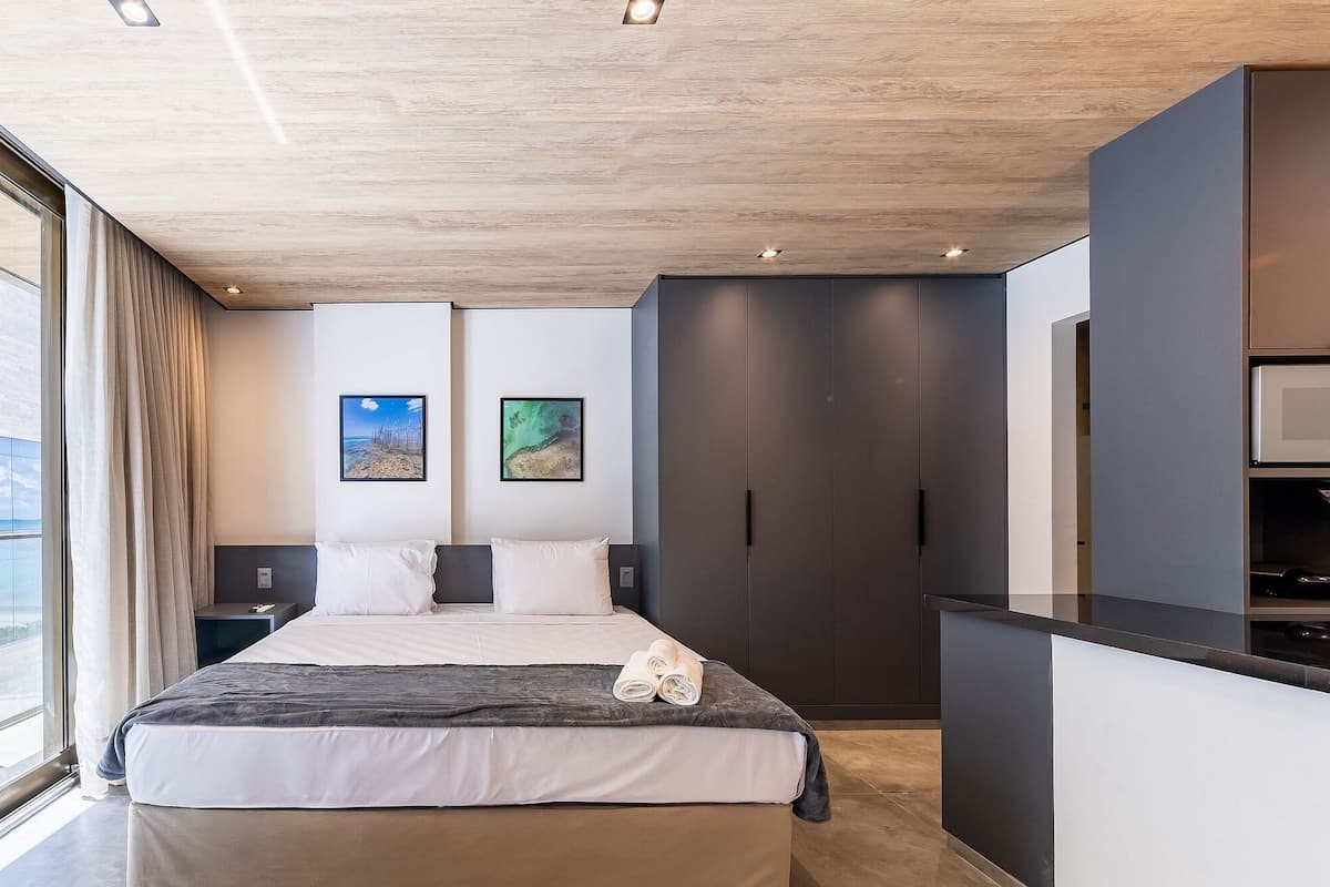 Une chambre d’hôtel moderne avec un grand lit, un bureau et une vue sur un immeuble, visible à travers de grandes fenêtres.