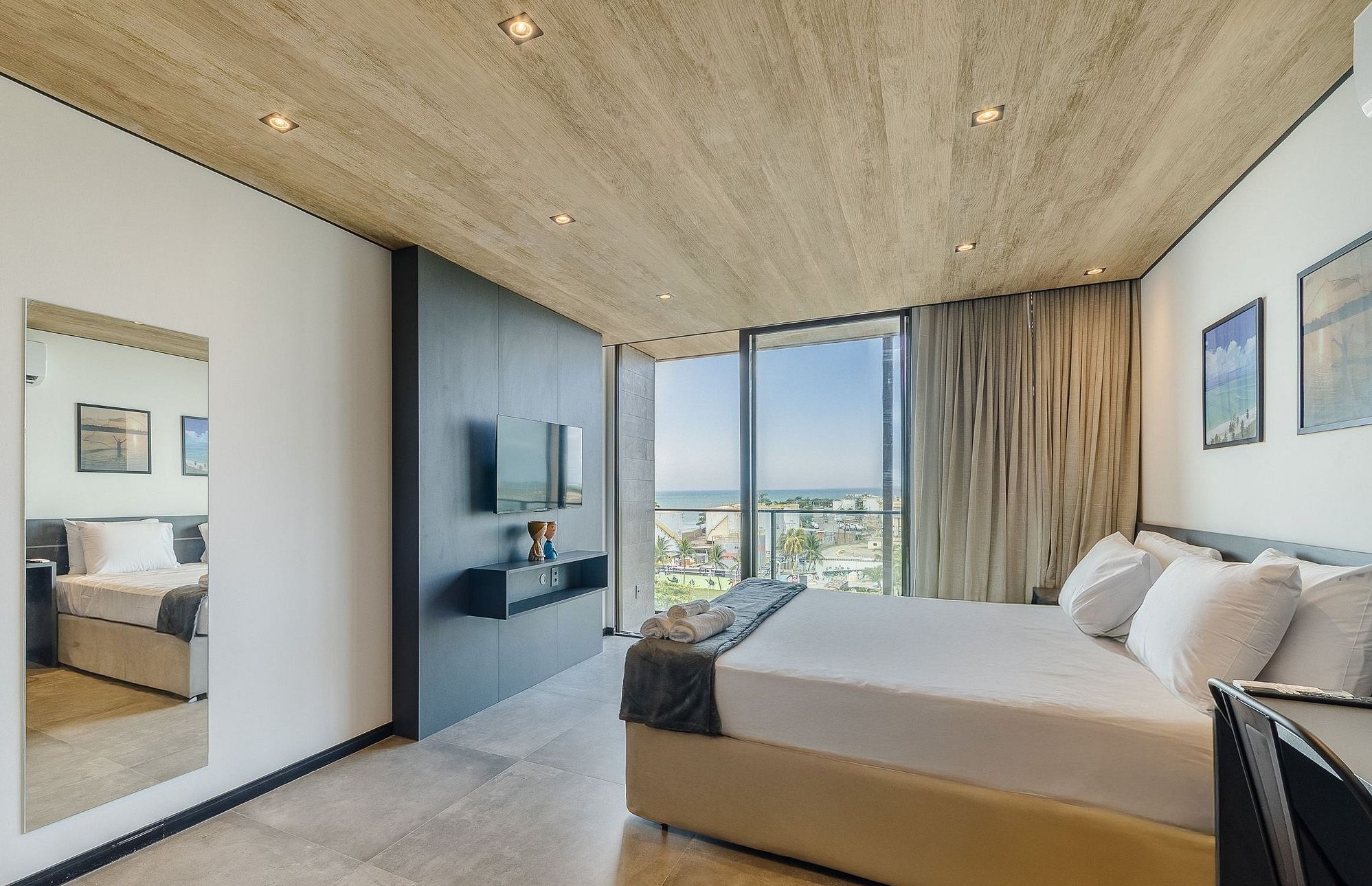 A modern hotel room with a large bed, a flat-screen TV, a view of the city, and a wooden ceiling.
