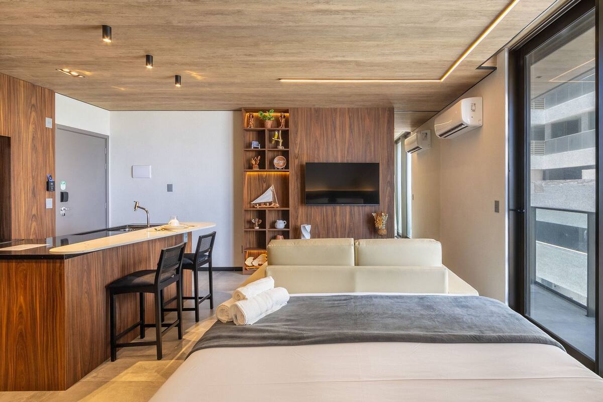 Une chambre d’hôtel moderne équipée d’un lit, d’un bureau avec des chaises, d’une télévision et d’un balcon offrant une vue sur la ville.