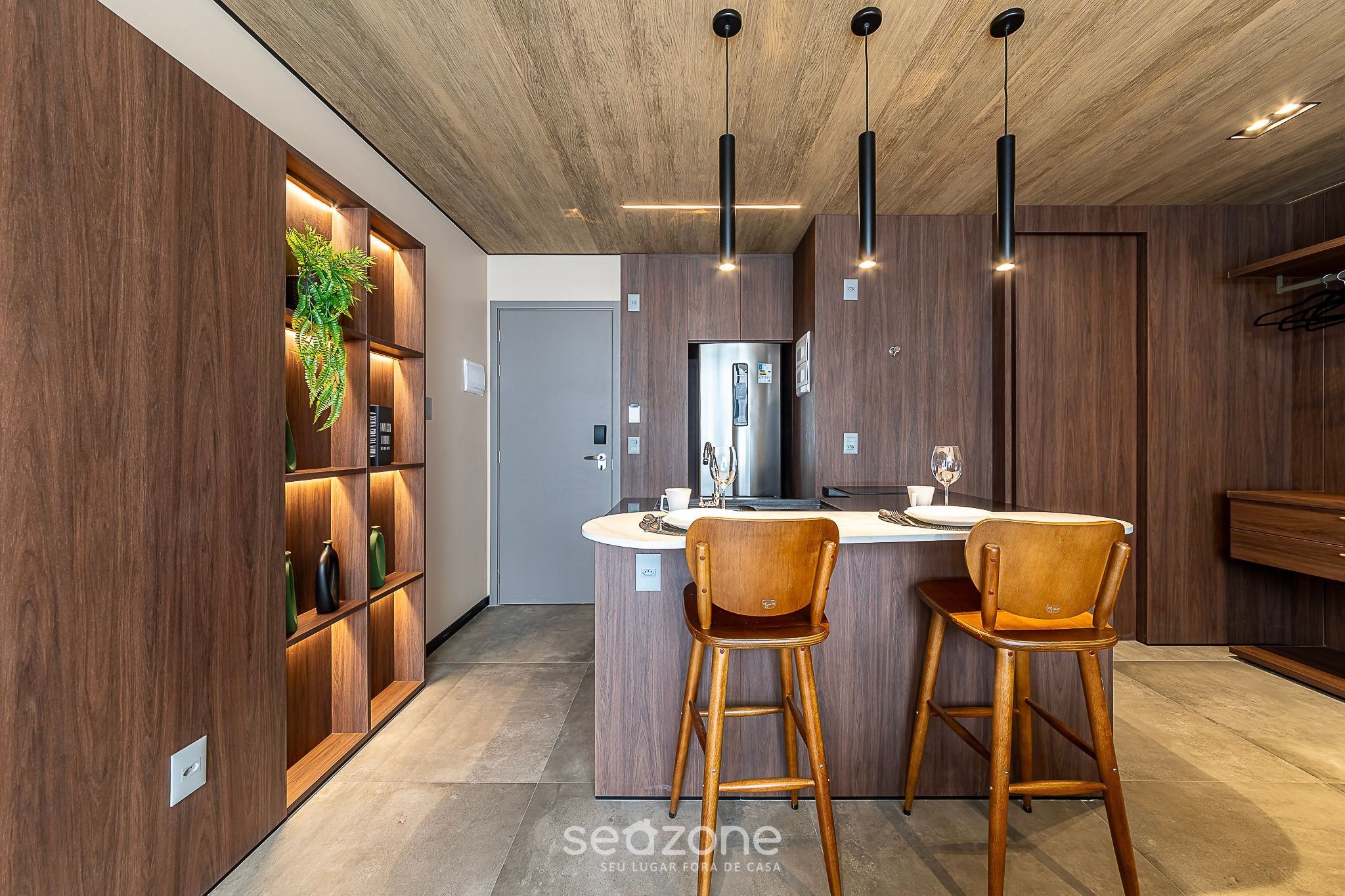 A modern kitchen with wooden cabinetry, a central island with bar stools, pendant lighting, and a visible refrigerator.