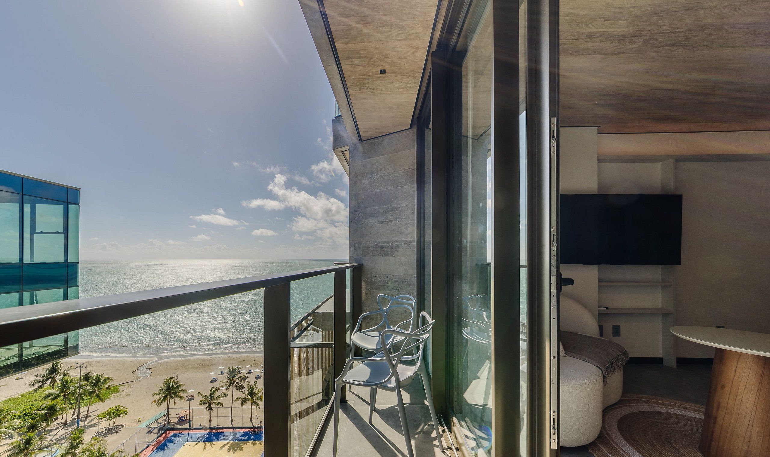 A balcony with a view of the beach and ocean, a pool, and a seating area with chairs and a table.