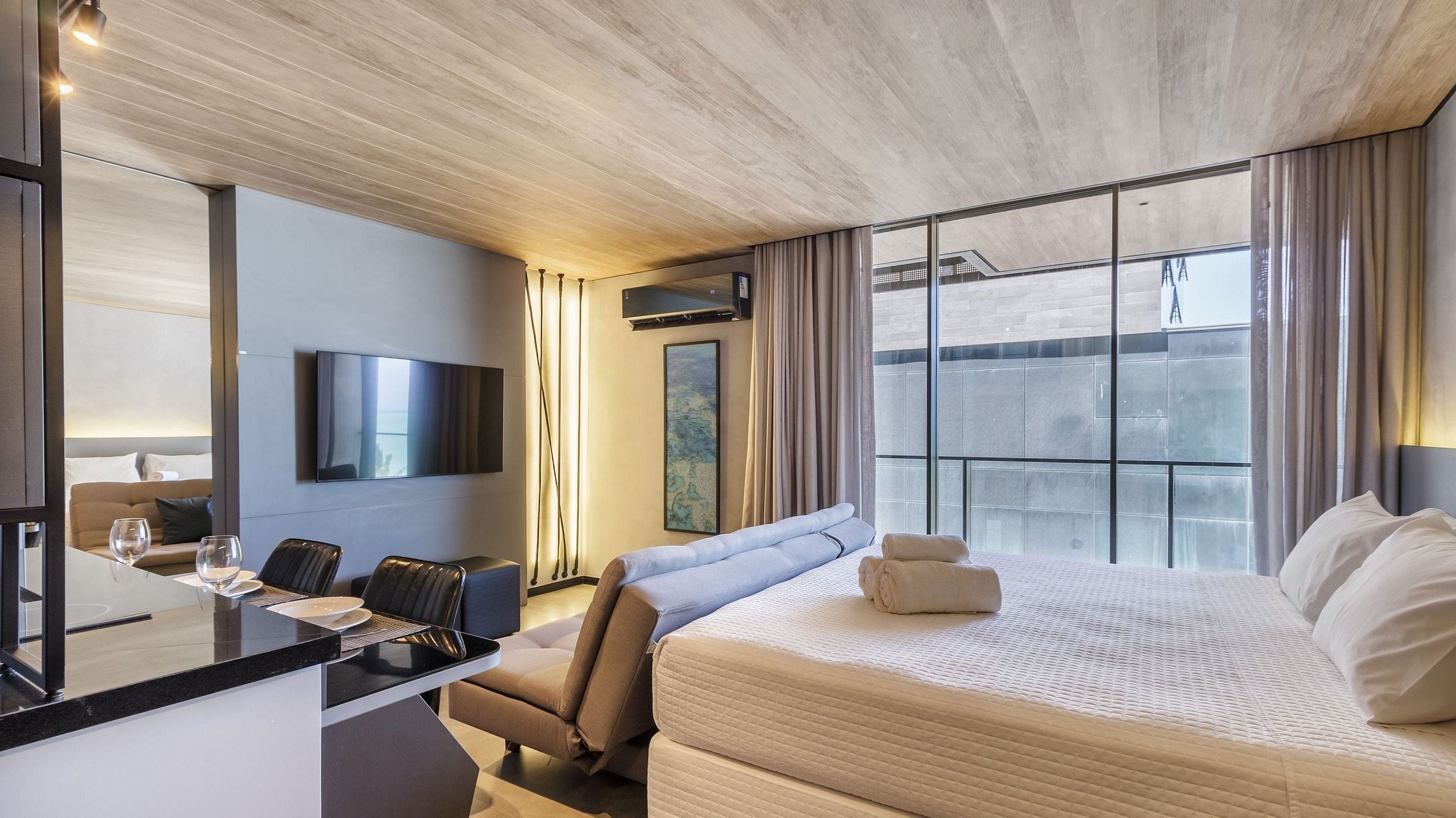 A hotel room with a bed, a desk with a chair, a flat-screen TV, and a large window with curtains.