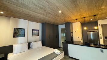 Un dormitorio moderno con cama, una kitchenette pequeña y un baño visible a través de una puerta abierta.