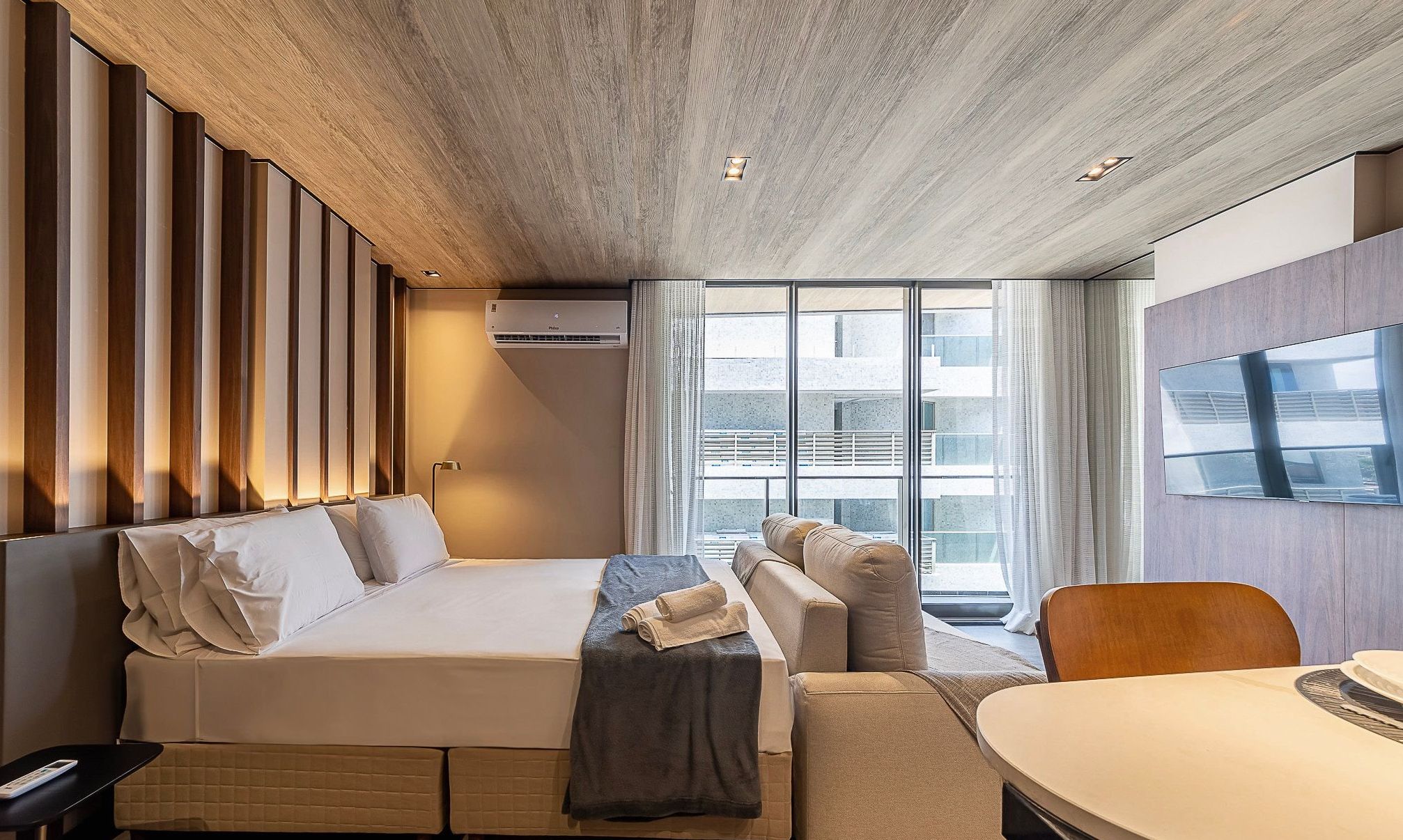 A hotel room with a bed, a sofa, a table, and a large window with a view of buildings.
