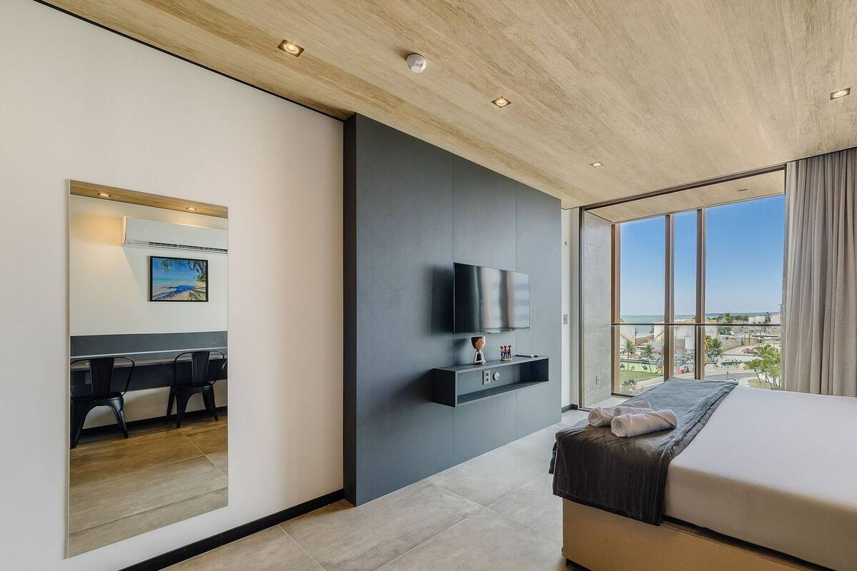 Une chambre d’hôtel moderne dotée d’un grand lit, d’un téléviseur à écran plat fixé au mur, d’un bureau avec des chaises et offrant une vue sur la plage grâce à de grandes fenêtres.