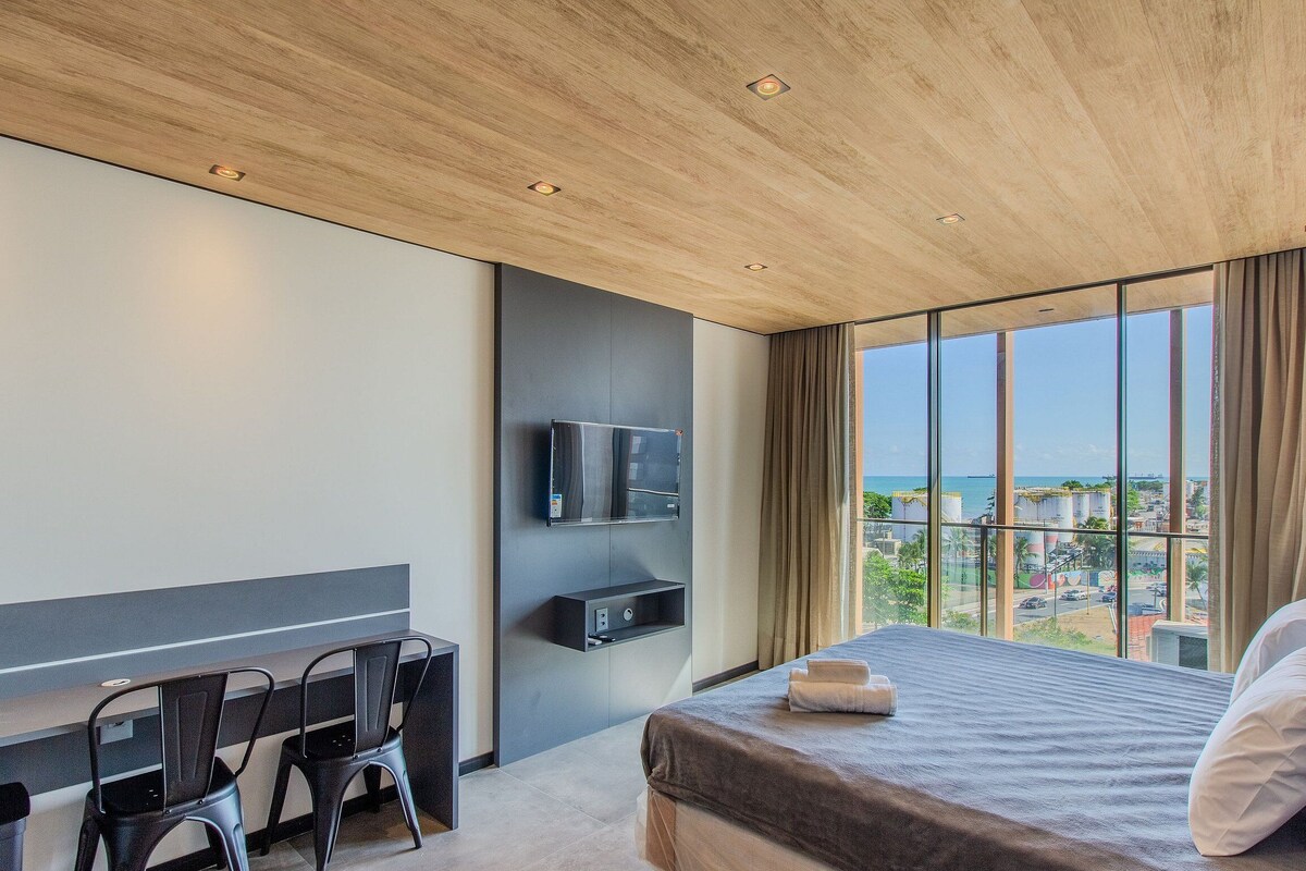 Ein modernes Hotelzimmer mit einem großen Bett, einem Schreibtisch mit Stuhl, einem an der Wand montierten Fernseher und Blick auf Stadt und Meer durch große Fenster.