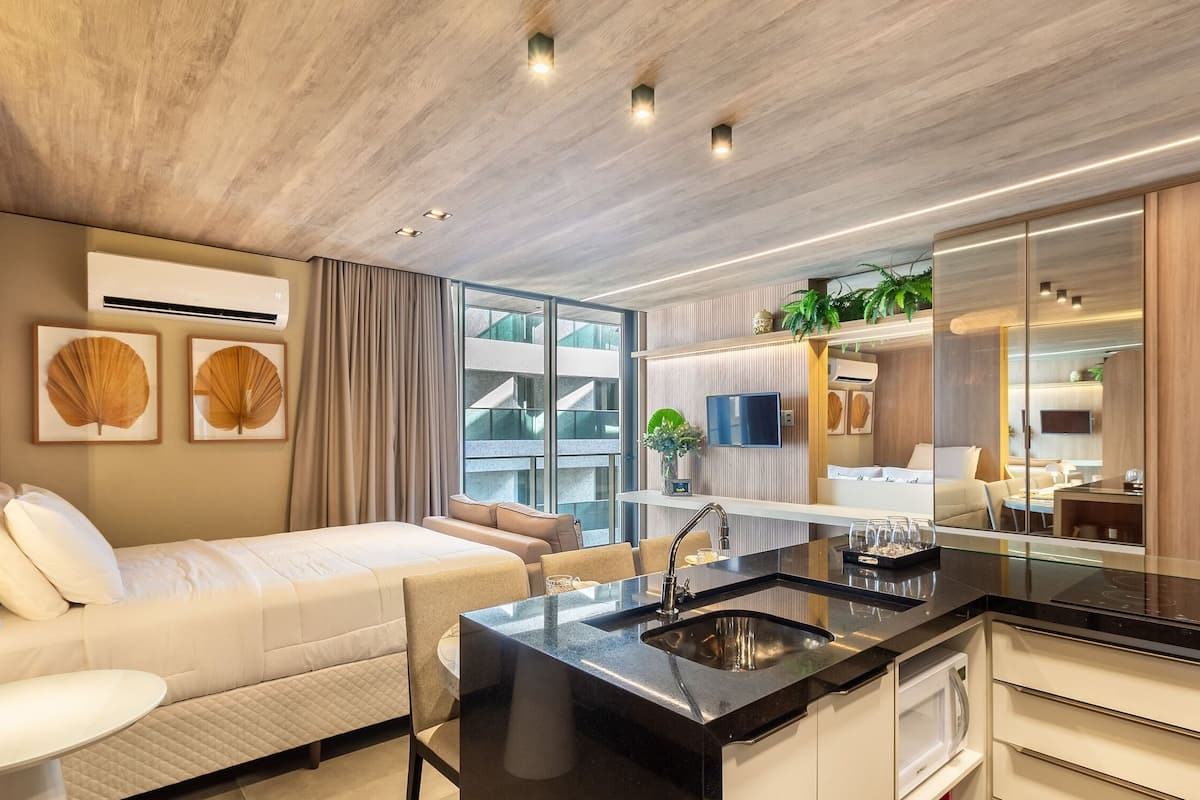 Une chambre d’hôtel moderne comprenant un lit, un petit coin salon, une cuisine avec un évier et un grand miroir.