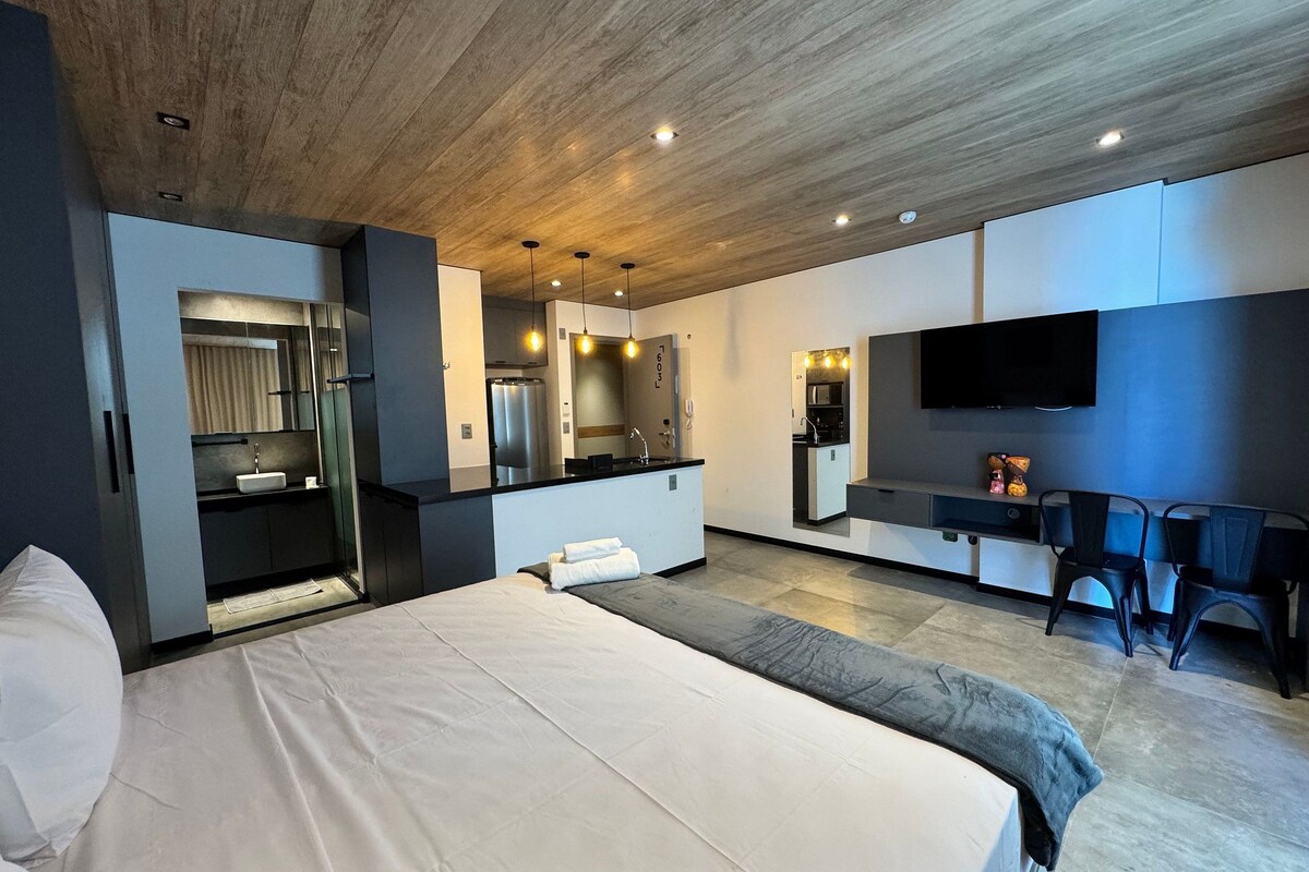 A modern hotel room with a large bed, a built-in desk, a kitchenette, and a TV mounted on the wall.