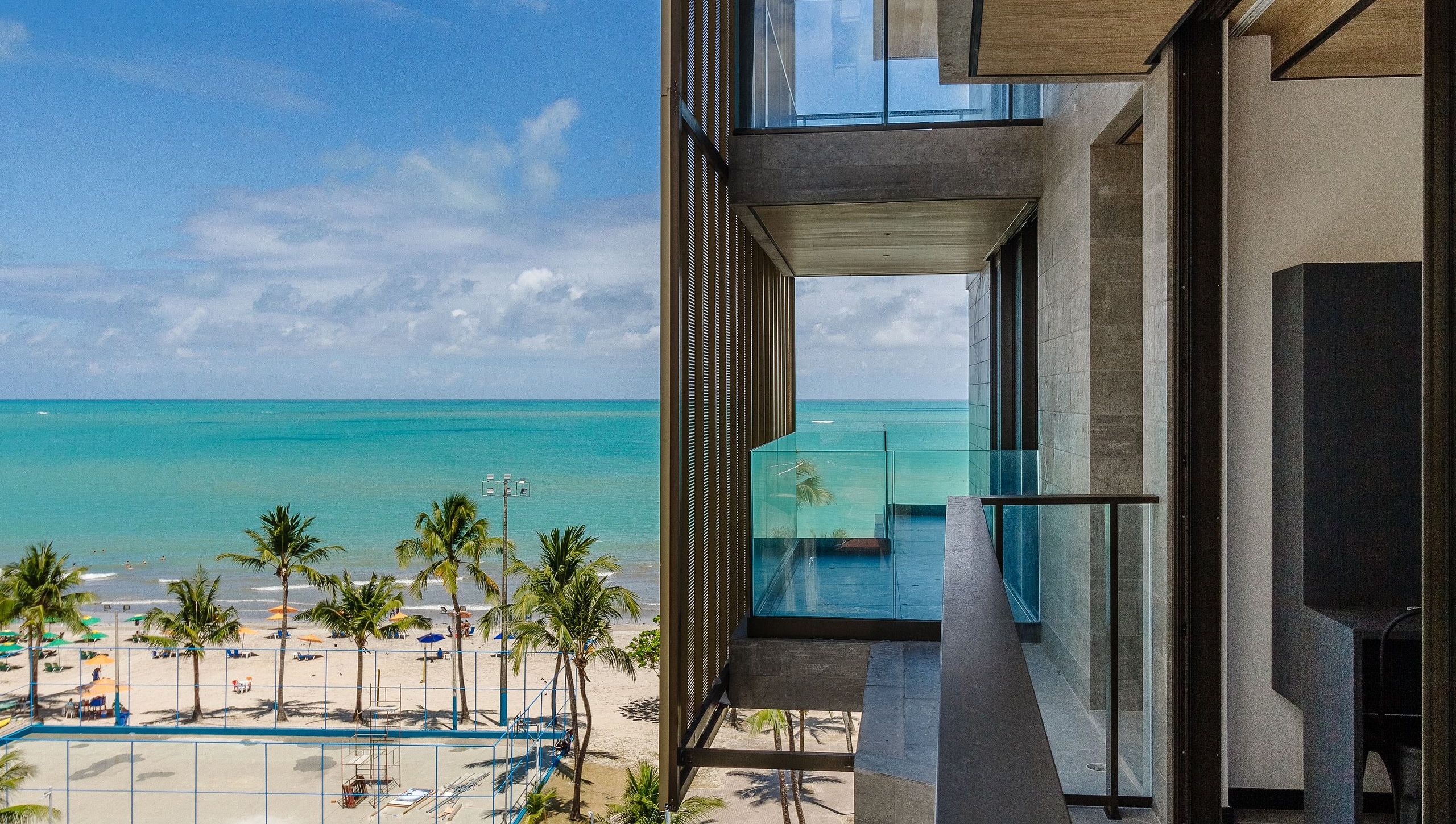 Un edificio moderno con un balcón de vidrio que da a una playa con palmeras y un mar azul cristalino.