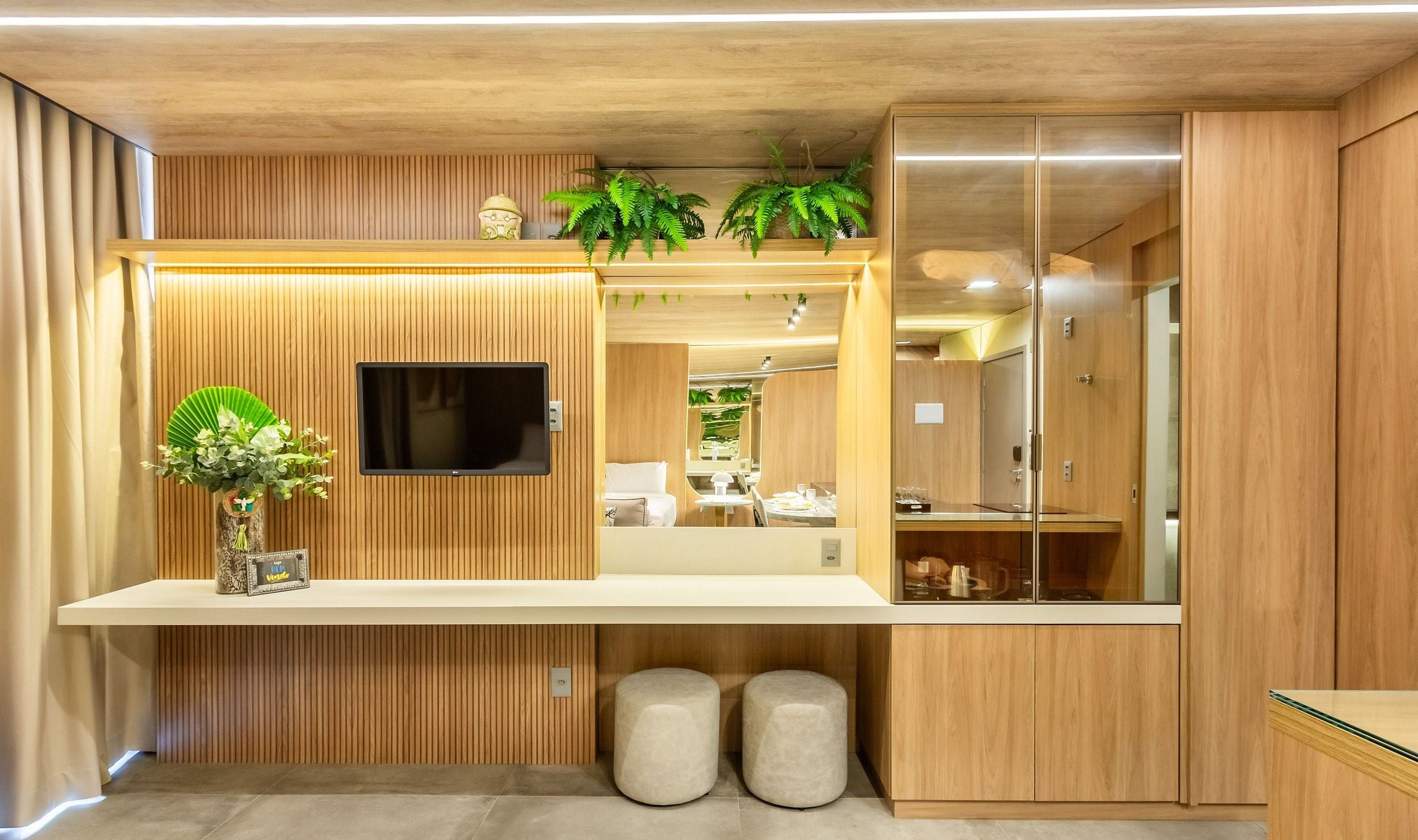 A modern hotel room with a wooden panel wall, a flat-screen TV mounted above a white console table, and a large mirror reflecting the room.