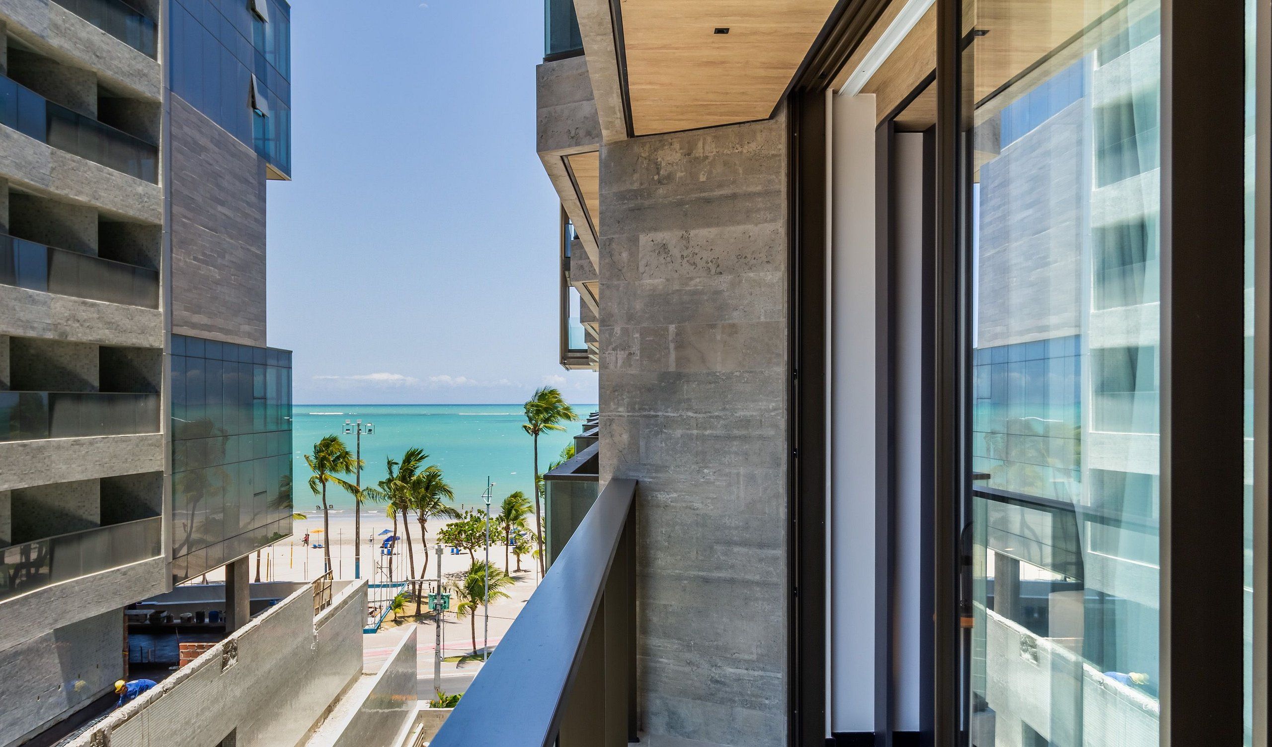 Ein modernes Gebäude mit einem Balkon mit Blick auf einen Strand, an dem Palmen und das Meer zu sehen sind.