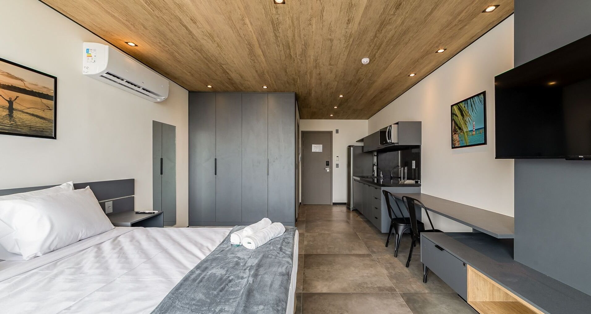 A modern hotel room with a bed, a desk, a kitchenette, and a TV.