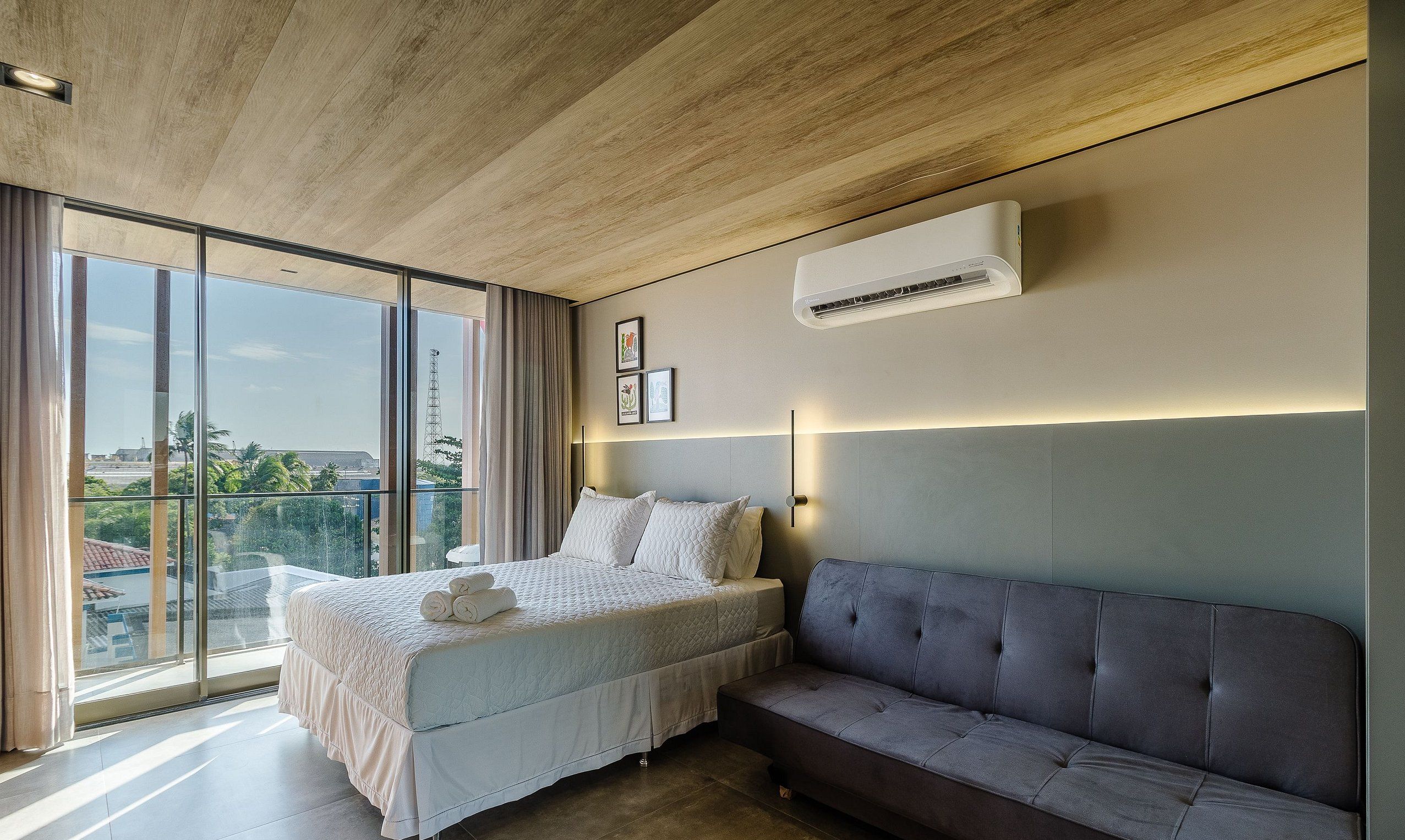 A modern bedroom with a large bed, a grey sofa, and a city view.
