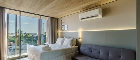 A modern bedroom with a large bed, a grey sofa, and a city view.