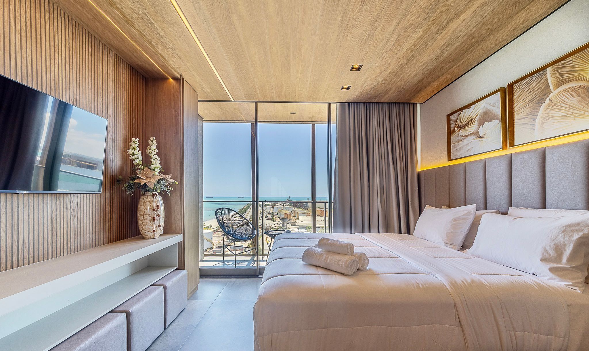 A modern hotel room with a large bed, a flat-screen TV, a balcony with a view of the sea, and a dining area with a chair.