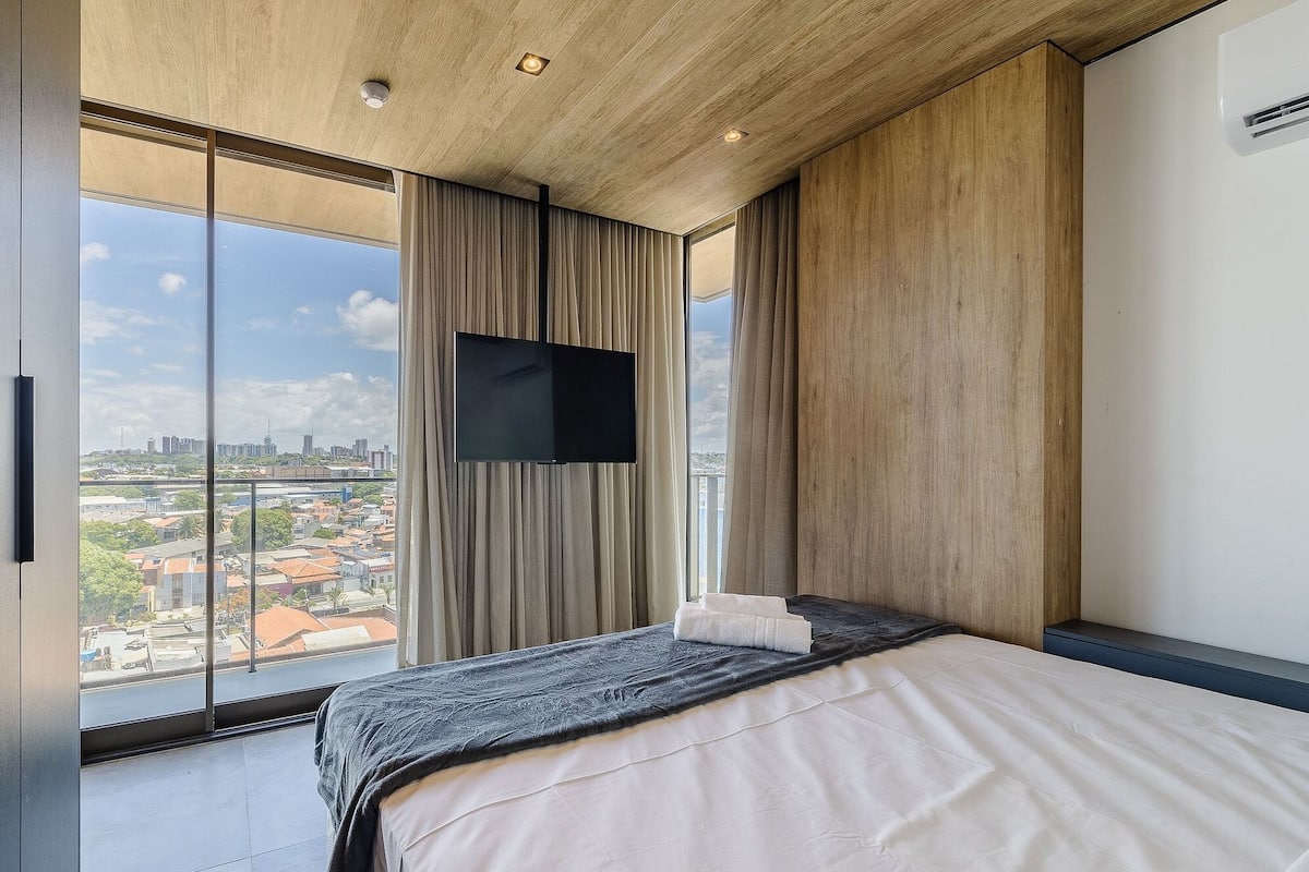 Une chambre d’hôtel moderne dotée d’un grand lit, d’un téléviseur à écran plat fixé au mur et offrant une vue sur la ville grâce à de grandes fenêtres du sol au plafond.
