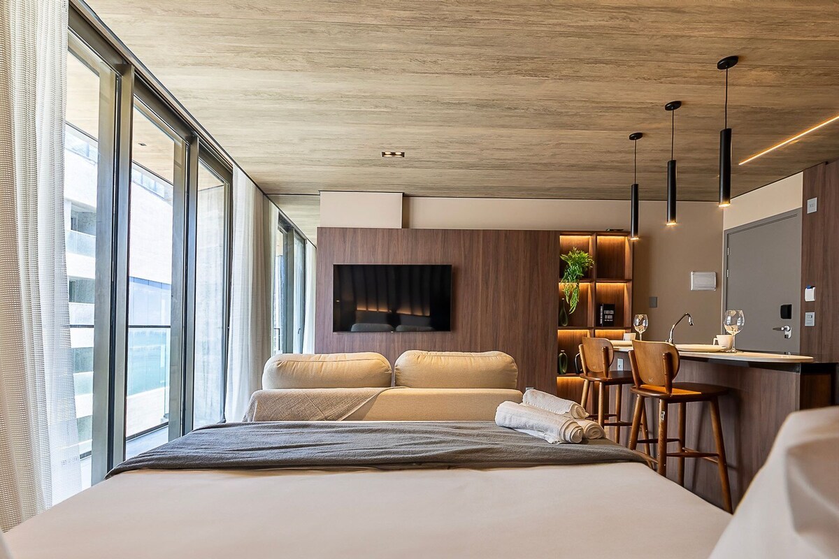 Ein modernes Hotelzimmer mit einem großen Bett, einem an der Wand montierten Fernseher, einer kleinen Küchenzeile mit einer Bar und großen Fenstern mit Blick auf die Stadt.