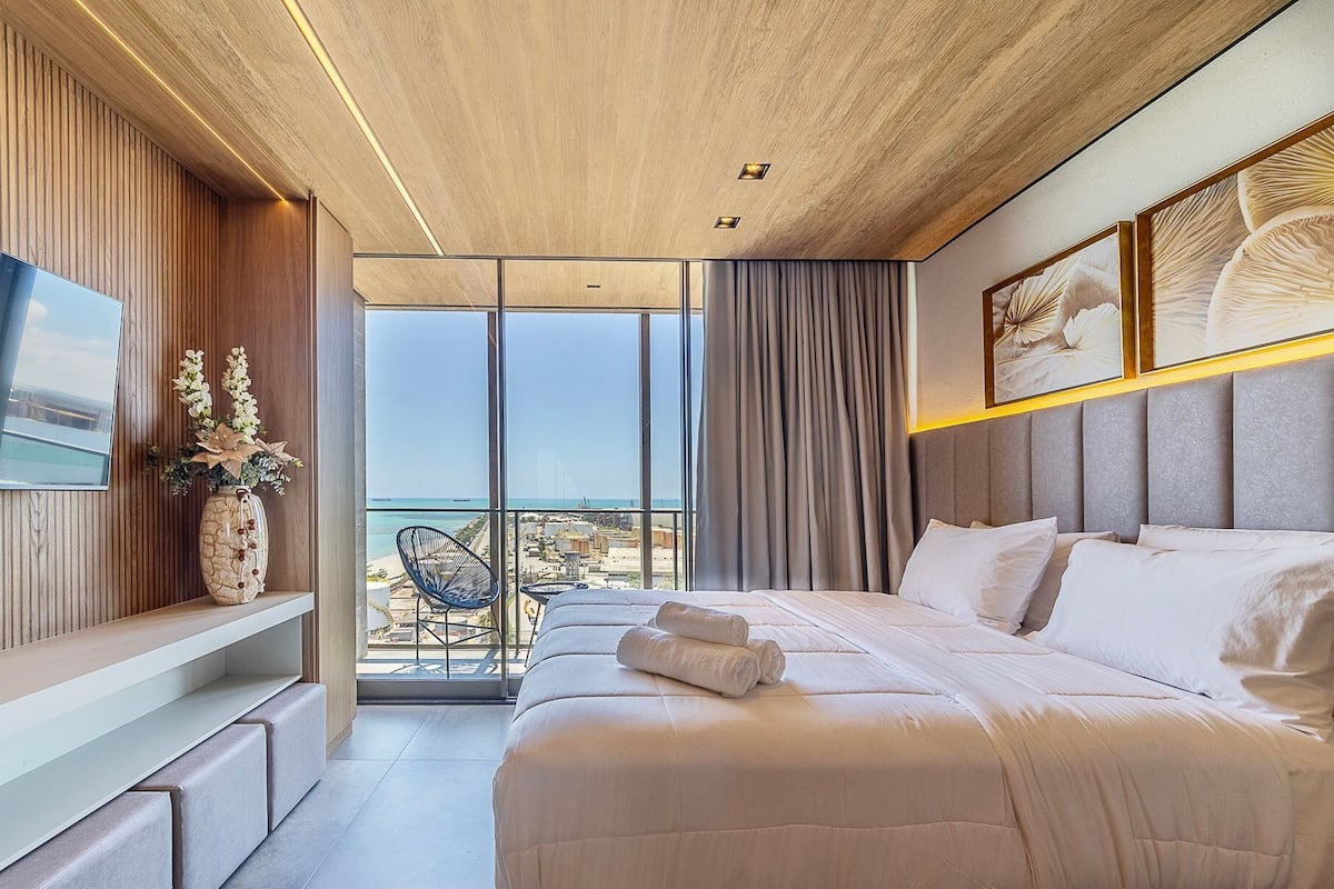 Une chambre d’hôtel moderne dotée d’un grand lit, d’une télévision et d’un balcon offrant une vue sur la plage, ainsi que d’un vase décoratif.