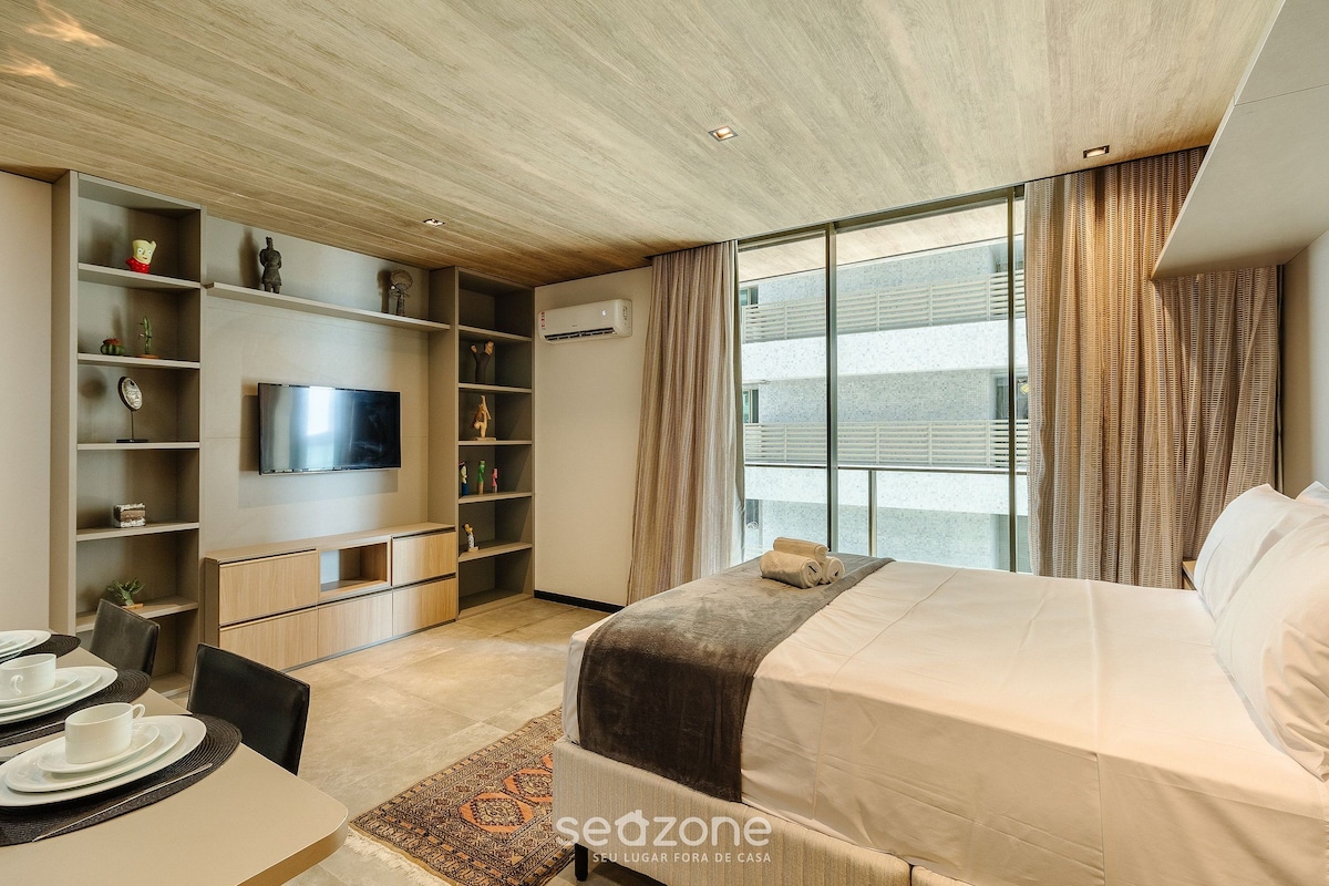 Habitación de hotel moderna con cama, televisor, zona de comedor y un gran ventanal con cortinas.