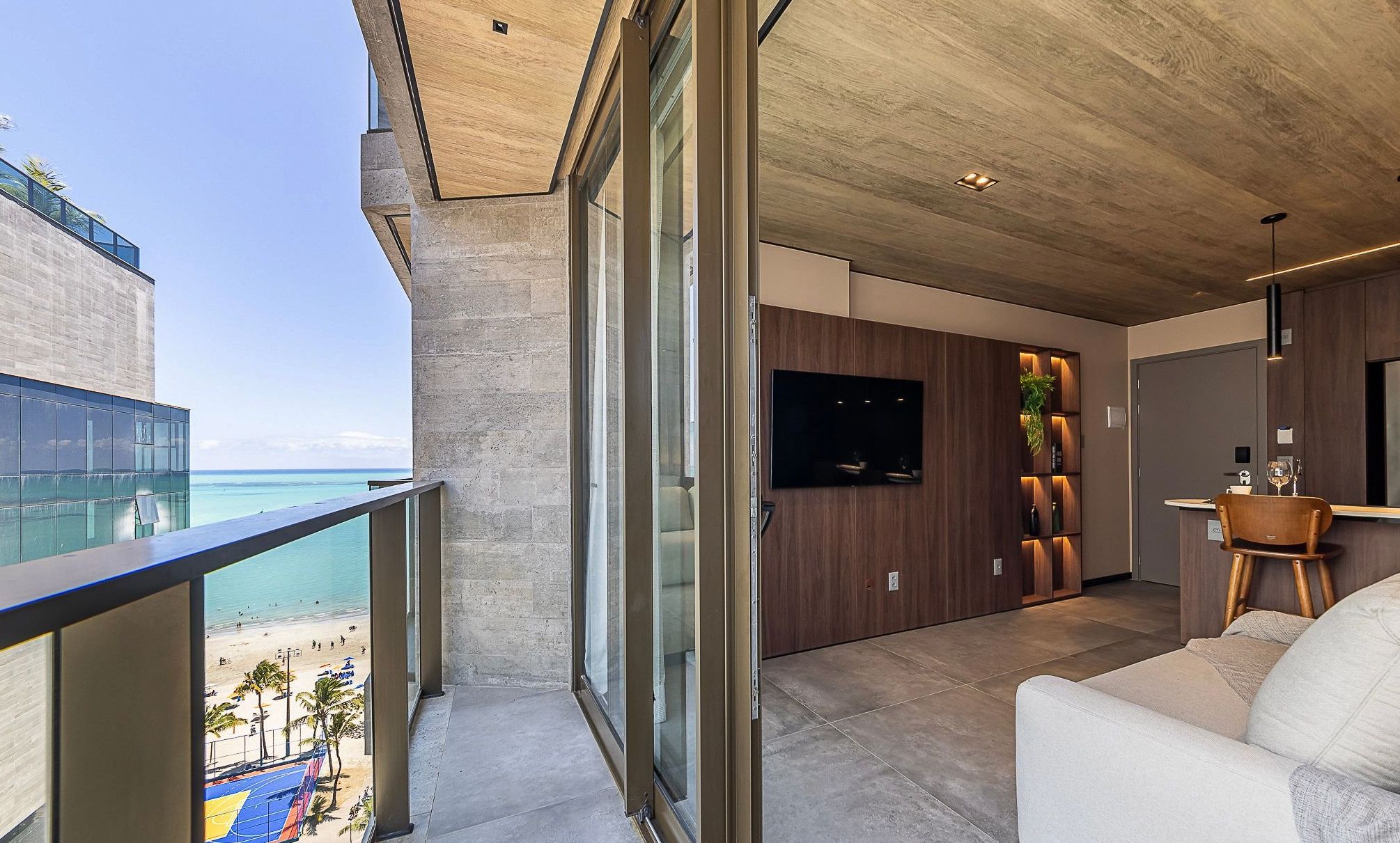Uma sala de estar moderna com uma televisão grande, um armário de madeira e uma varanda com vista para a praia.