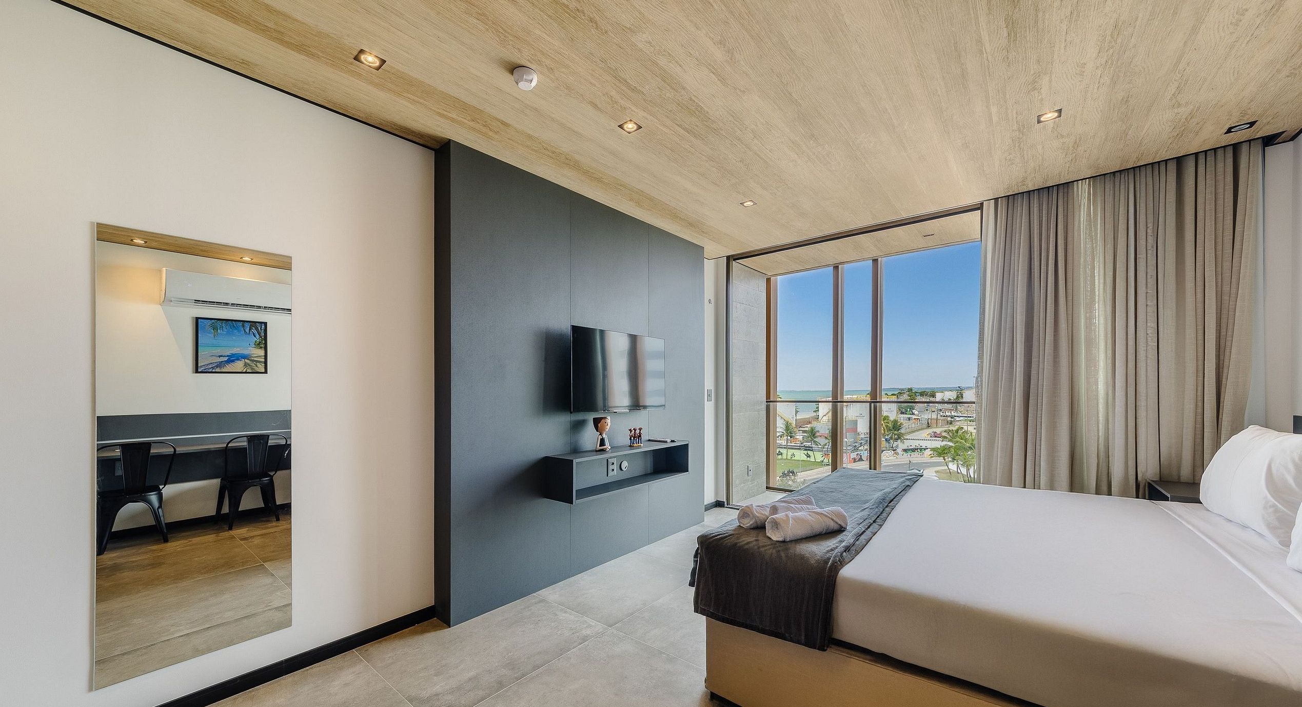 Una habitación de hotel moderna con una cama grande, un televisor montado en la pared, un escritorio con una silla y vistas a la ciudad a través de amplios ventanales.