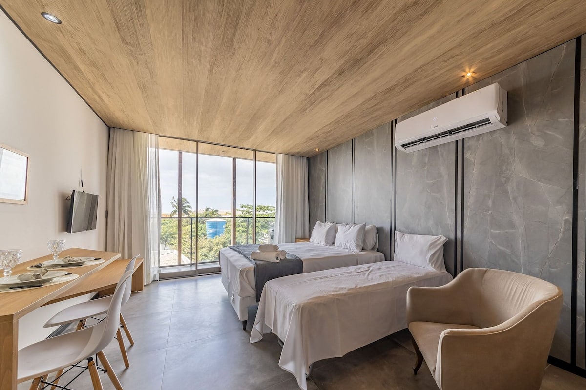 Une chambre d’hôtel moderne avec un plafond en bois, un lit, un bureau avec des chaises, une télévision et une grande fenêtre donnant sur un paysage verdoyant.