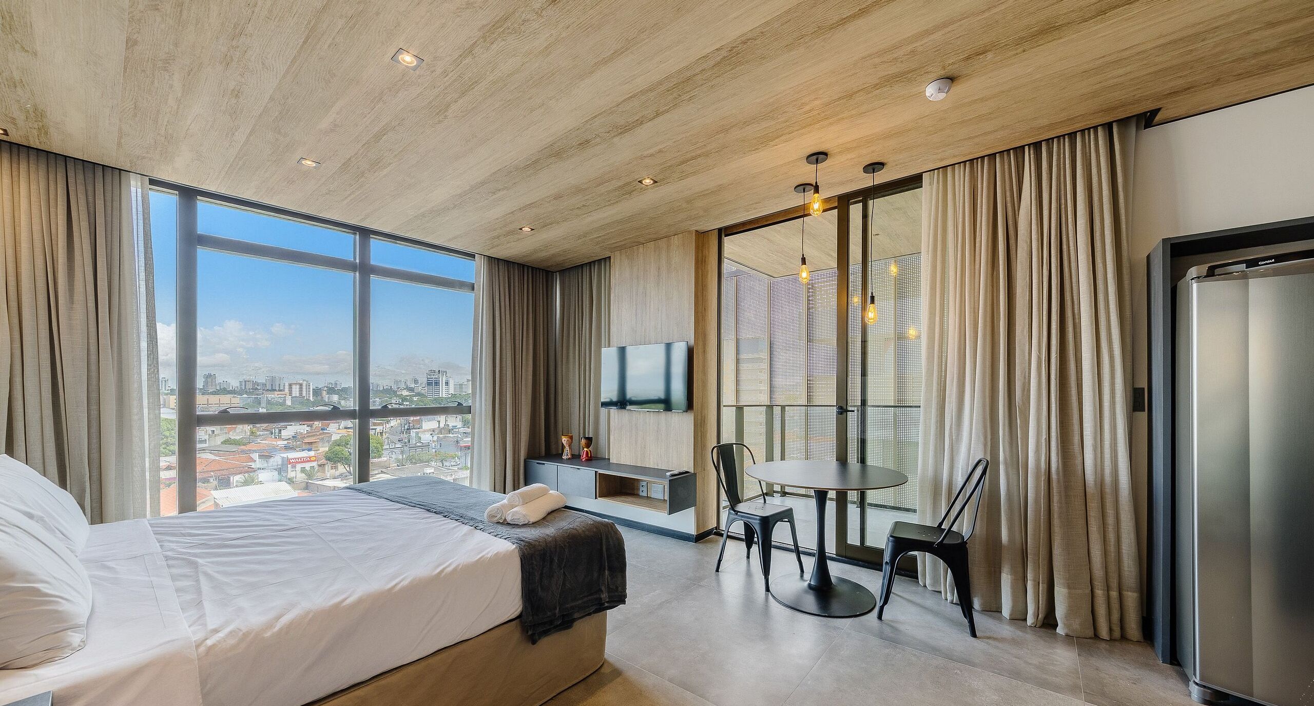 Una habitación de hotel moderna con una cama grande, una mesa de comedor con sillas, un televisor montado en la pared y vistas de la ciudad a través de amplios ventanales.