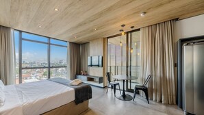 Ein modernes Hotelzimmer mit einem großen Bett, einem Esstisch mit Stühlen, einem an der Wand befestigten Fernseher und Blick auf die Stadt durch große Fenster.