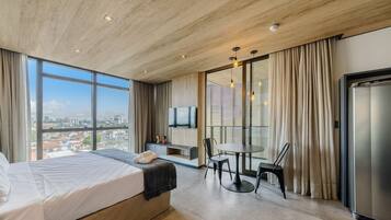 Una habitación de hotel moderna con una cama grande, una mesa de comedor con sillas, un televisor montado en la pared y vistas de la ciudad a través de amplios ventanales.