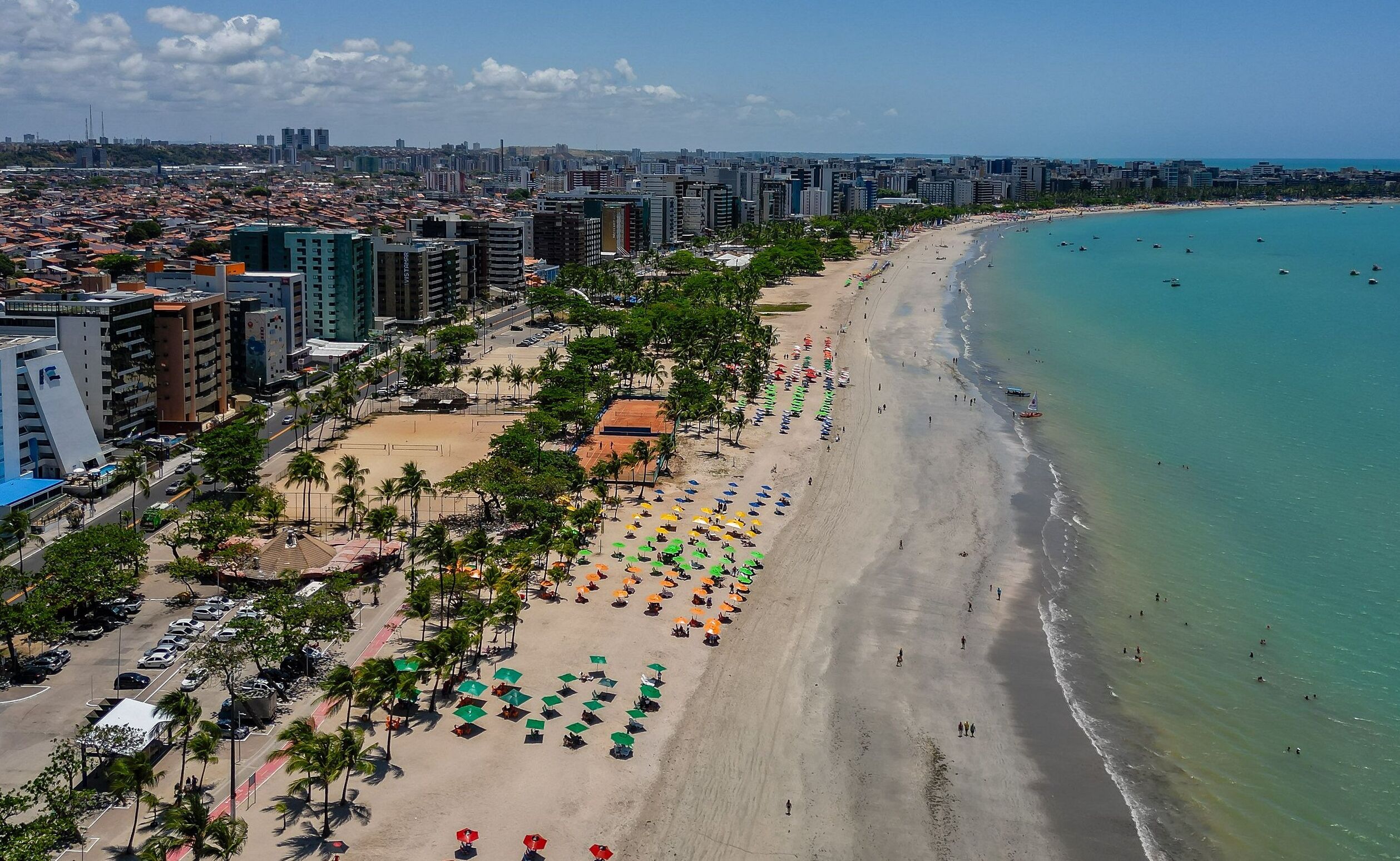 Paisagem costeira urbana com uma praia repleta de guarda-sóis e palmeiras.