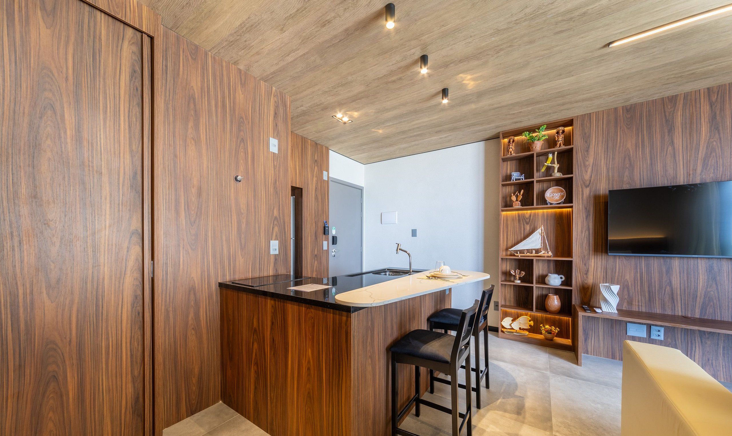 Eine moderne Küche mit Holzschränken, einer Mittelinsel, Barhocker, einem integrierten Fernseher und einem Regal mit Dekoration.