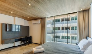 A modern hotel room with a large bed, a desk, a flat-screen TV, and a view of the cityscape through floor-to-ceiling windows. - NWTs - Estúdios com Vista Lateral Mar (Maceió)