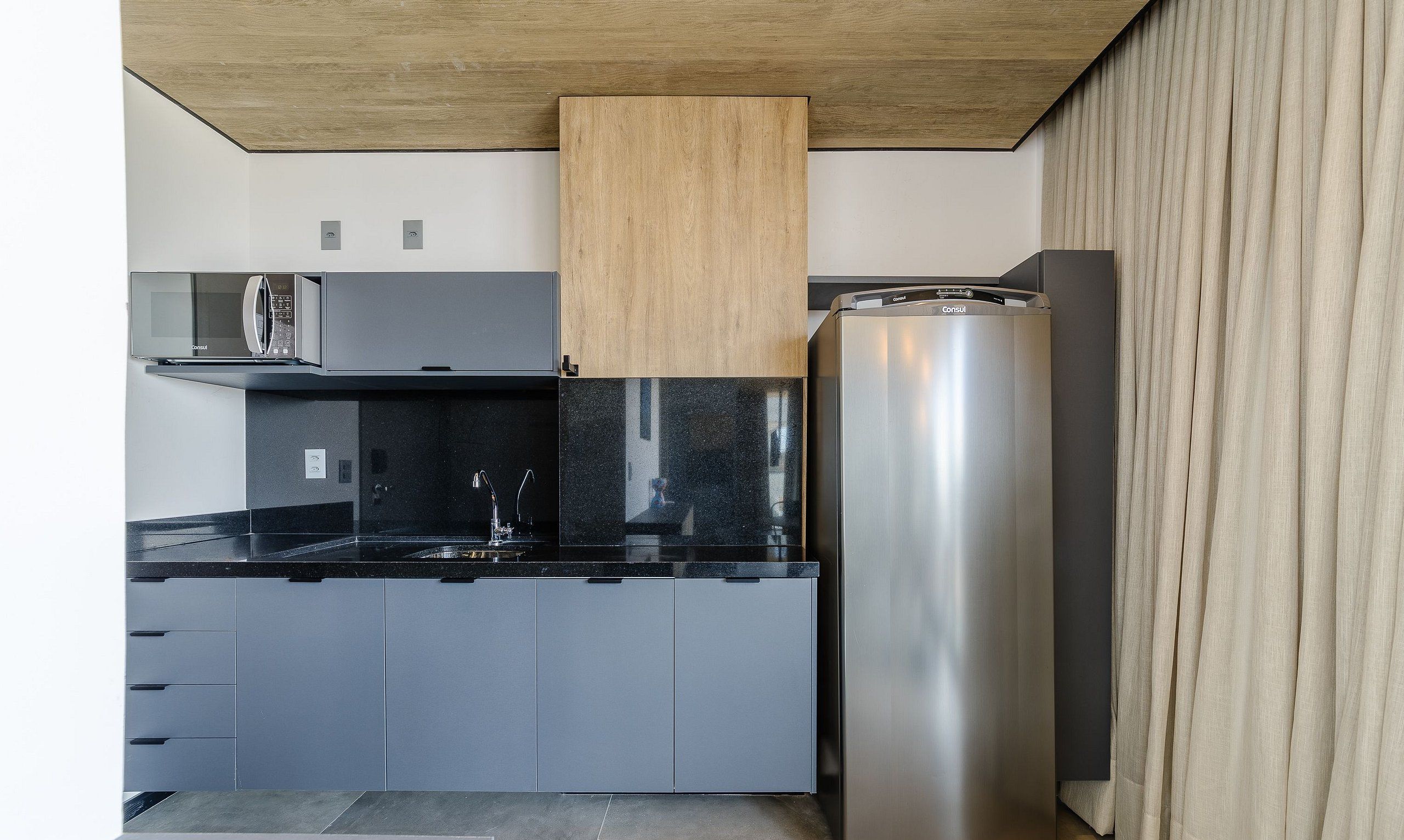Une cuisine moderne avec des meubles gris, un plan de travail noir, un réfrigérateur en acier inoxydable et un plafond en bois.